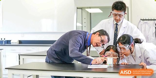 A teacher, possibly a science instructor, leans over a desk to assist students with their written work in a classroom at Sedbergh School Fuzhou.