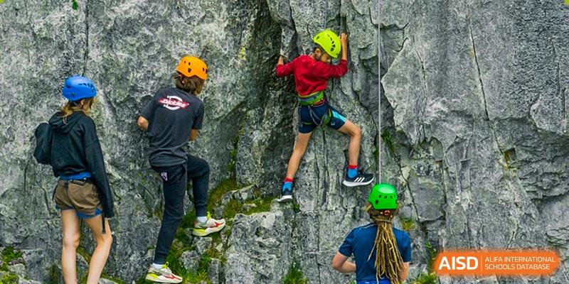 Students wearing helmets and safety harnesses rock climbing outdoors on a natural-looking cliff face, showcasing an adventure sport activity at Sedbergh School Fuzhou.
