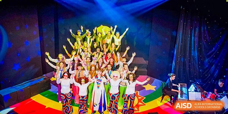 A large group of students in colorful costumes posing with their hands raised on a brightly lit stage after a performance at Sedbergh School Fuzhou.