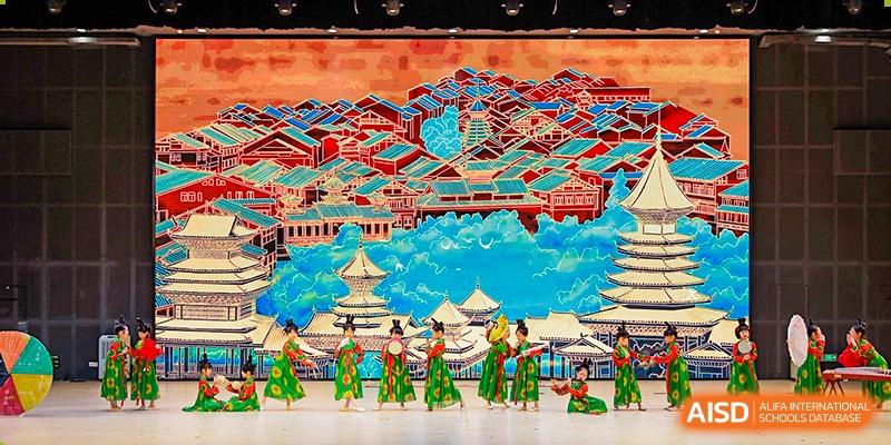 Students performing a traditional Chinese dance in elaborate costumes on a stage set against a detailed cultural backdrop at Sedbergh School Fuzhou.