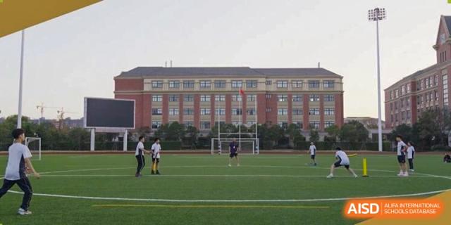 Students playing soccer on a wide green sports pitch in front of the impressive red brick school main building at Sedbergh School Fuzhou.