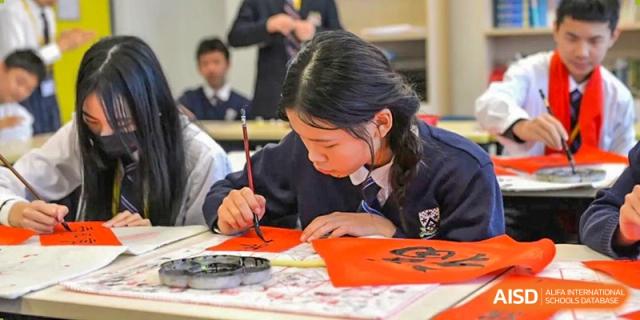 A female student from Dulwich International High School Programme Hengqin (DHHQ) practicing Chinese calligraphy with a brush, creating characters on a piece of red paper.