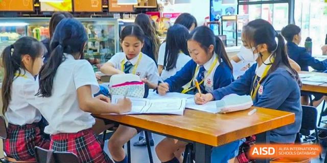 A group of female students from Dulwich International High School Programme Hengqin (DHHQ) focused on writing or completing assignments at a table in a bright, casual setting.