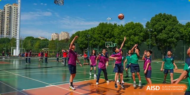 A group of enthusiastic students from Dulwich International High School Programme Hengqin (DHHQ) playing basketball on an outdoor court under a bright blue sky.
