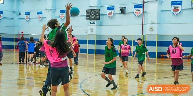 Female students from Dulwich International High School Programme Hengqin (DHHQ) practicing or playing basketball indoors, with one student attempting a jump shot.