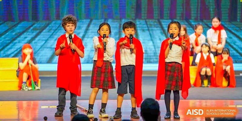 A group of young students from Dulwich International High School Programme Hengqin (DHHQ) in matching red and plaid uniforms singing into microphones on a stage.