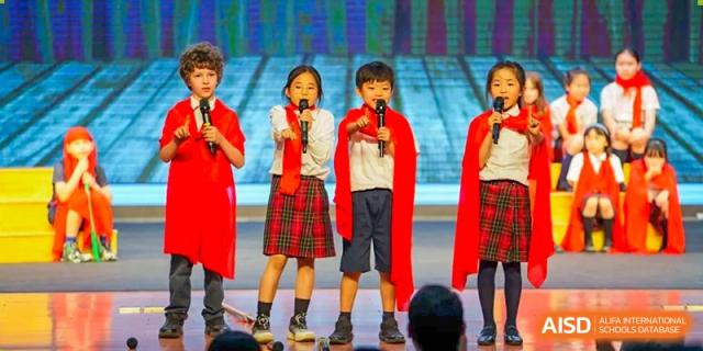 A group of young students from Dulwich International High School Programme Hengqin (DHHQ) in matching red and plaid uniforms singing into microphones on a stage.