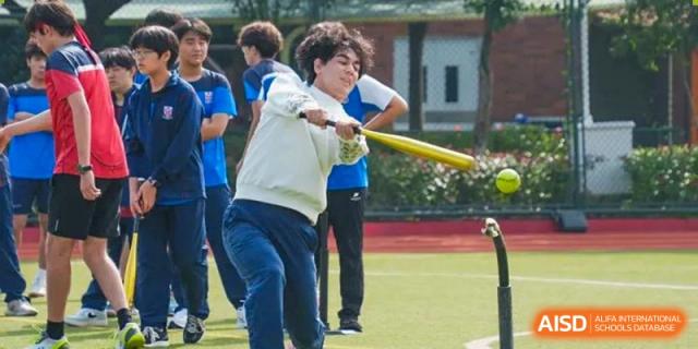 A male student from Dulwich International High School Programme Hengqin (DHHQ) hitting a ball with a bat while his classmates watch, likely during a softball or baseball practice.