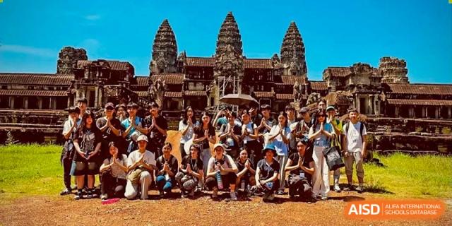A large group of students and teachers from Dulwich International High School Programme Hengqin (DHHQ) posing for a group photo in front of a historical temple or ruins (possibly Angkor Wat) during a school trip.
