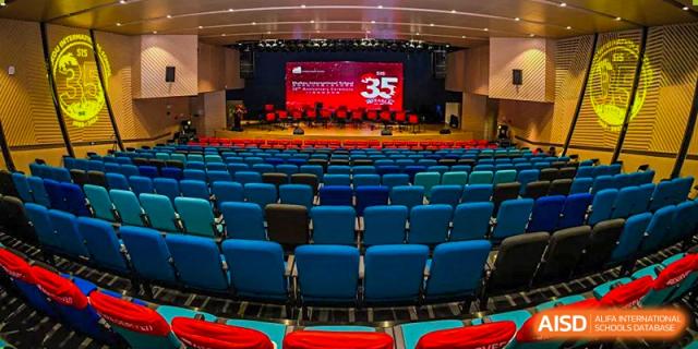The interior of the large, modern auditorium at Shekou International School, featuring rows of blue seats and a stage banner.