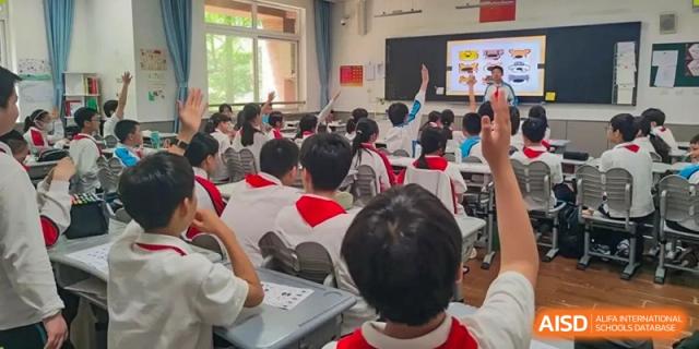 A student raising a hand to answer a question or participate, with other students seated at desks in the classroom at Shanghai Nanyang Model Private School.