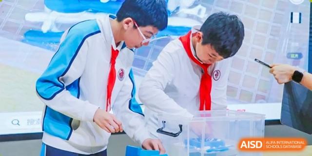 Two students in white lab coats or shirts with red scarves collaborating on a science experiment or project using plastic containers at Shanghai Nanyang Model Private School.
