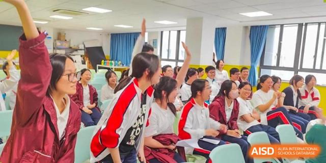 A group of students in red and white uniforms enthusiastically raising their hands to participate in a classroom discussion at Shanghai Nanyang Model Private School.
