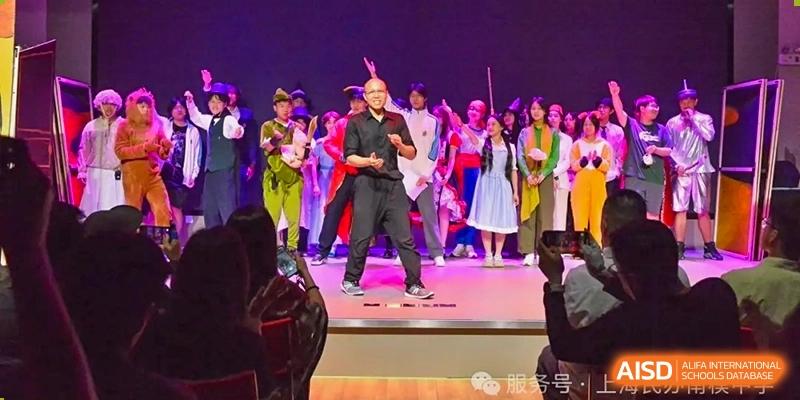A performer on a stage in black clothes, gesturing to an audience with a crowd of people dressed in costume behind him at Shanghai Nanyang Model Private School.