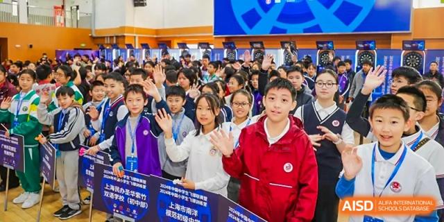 Students waving enthusiastically towards the camera in a large indoor hall or auditorium at Shanghai Nanyang Model Private School.