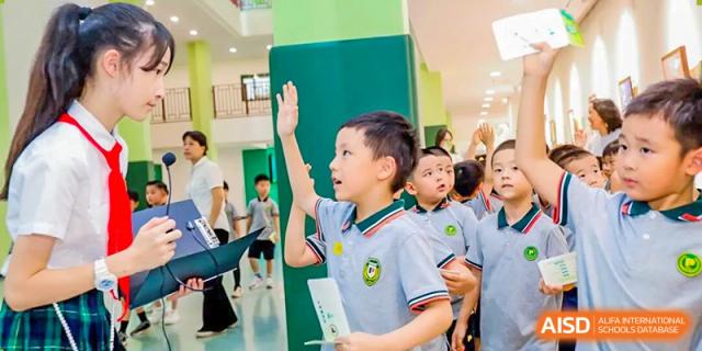 An enthusiastic young Shanghai Pinghe School student in a polo uniform raises his hand high in a bright school hallway or event.