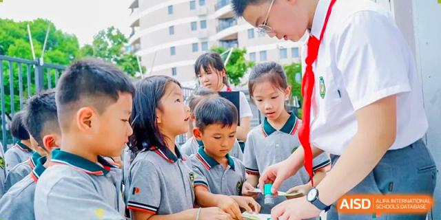 A Shanghai Pinghe School teacher in a red tie oversees a small group of young students engaged in a collaborative, hands-on activity outdoors.