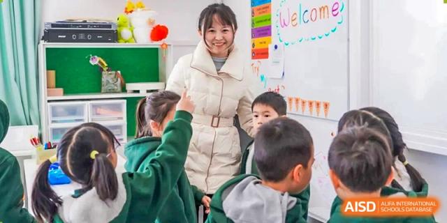 A Shanghai Pinghe School teacher smiles at her young students, who are eagerly raising their hands in a bright, decorated classroom.