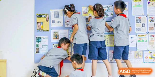 A group of young Shanghai Pinghe School students, some older and some younger, work collaboratively on a project near a bulletin board.