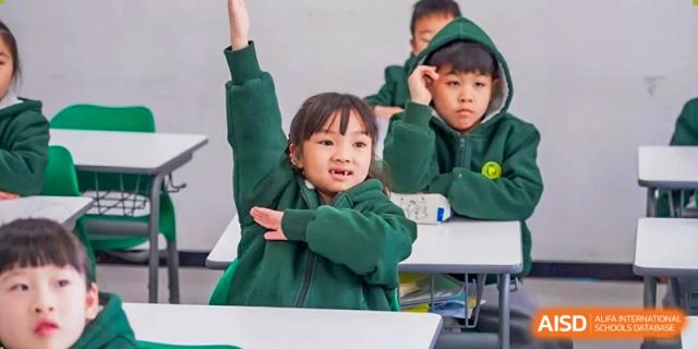 A Shanghai Pinghe School primary student in a green sweater raises her hand high while sitting at a desk with other classmates.