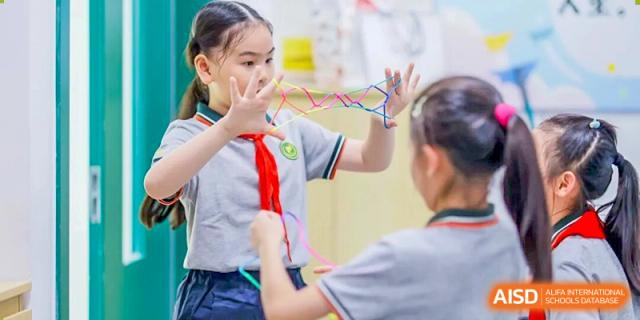 Two young Shanghai Pinghe School students in uniform and red scarves working together on a colorful string or craft project.