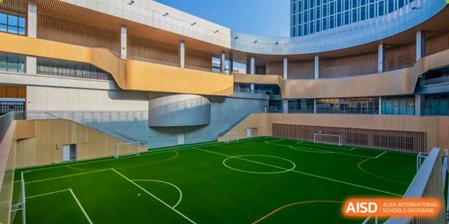Exterior view of a school building with modern, curved architecture, featuring an outdoor sports field with artificial turf in the foreground at Shanghai Pinghe School.