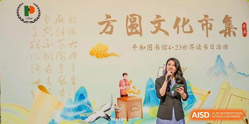 A teacher or speaker holding a microphone and addressing an audience in front of a colorful banner or backdrop with Chinese text at Shanghai Pinghe School.
