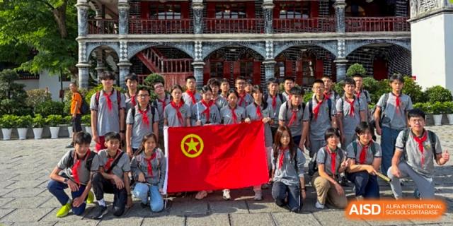 A large group of students in uniform and red scarves holding a red and yellow flag and posing outdoors in front of a traditional-style building at Shanghai Pinghe School.
