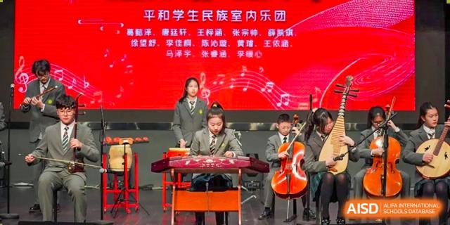 Students performing a Chinese orchestral piece with traditional instruments on a brightly lit stage with red backdrops at Shanghai Pinghe School.