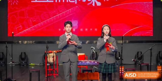 Two student hosts, a boy and a girl, in school uniform standing on a stage with a bright red backdrop, leading an event at Shanghai Pinghe School.