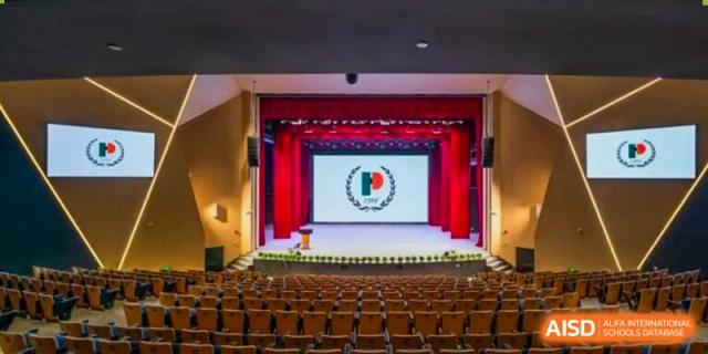 A full view of an auditorium stage with red curtains and empty tiered seating, featuring the school crest on the backdrop at Shanghai Pinghe School.