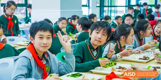 A group of smiling Shanghai Pinghe School students in green sweaters and uniforms eating together in the dining hall or classroom, possibly having a healthy lunch.