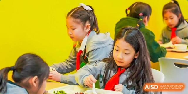 Two young female Shanghai Pinghe School students in uniform and red scarves focusing on their food during a meal, suggesting a health and nutrition focus.