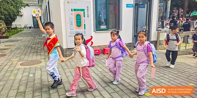 Three young female students from Beijing Bayi School International Department walk together holding hands, carrying backpacks, and smiling as they enter the school grounds.