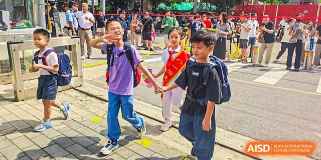 Two young students from Beijing Bayi School International Department, a boy with a large backpack and a girl wearing a red sash, walk across a crosswalk while holding hands.