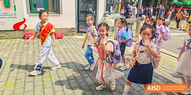 A group of young female students from Beijing Bayi School International Department, wearing bright clothes and backpacks, walk together on a paved path outside the school building.