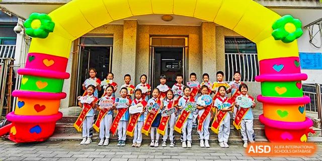 A large group of students from Beijing Bayi School International Department are lined up in front of a yellow school building, wearing matching red, white, and blue uniforms with sashes for a group photo.