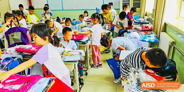 A busy classroom scene at Beijing Bayi School International Department with many young students seated at desks, some interacting with each other and an adult near the foreground.