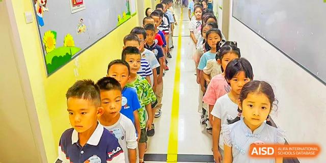 A long line of young students in a bright hallway at Beijing Bayi School International Department, standing neatly behind a yellow line as they wait their turn.
