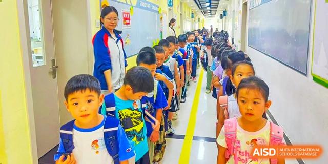 A teacher standing with a line of elementary students waiting in a school corridor at Beijing Bayi School International Department. The students are looking toward the camera.
