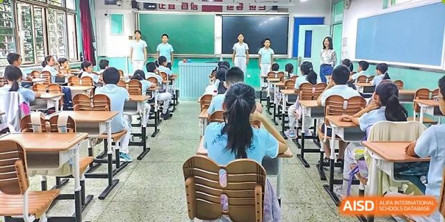A wide view of a classroom at Beijing Bayi School International Department where students are seated at desks, looking toward the front where two teachers stand near the chalkboard.