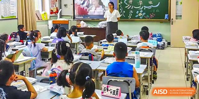 A classroom scene at Beijing Bayi School International Department showing a teacher at the front demonstrating a lesson on a large screen monitor to the seated students.