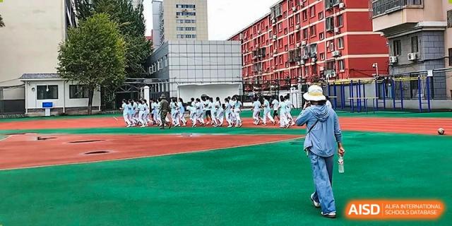 Students in uniforms practicing a routine or physical activity on a red and green outdoor sports ground at Beijing Bayi School International Department, with a large residential building in the background.