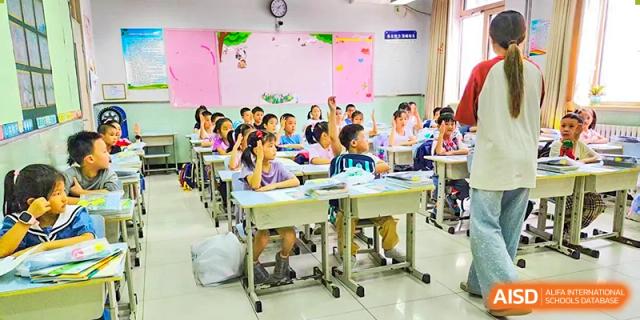 A lively classroom at Beijing Bayi School International Department where many students are sitting at their desks with their hands raised, ready to answer a question.