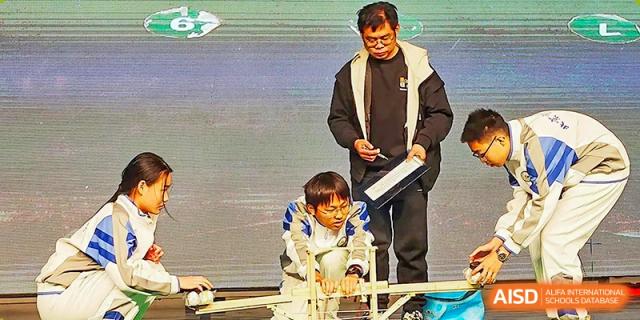 A teacher wearing glasses and a dark jacket helps a kneeling student on a stage with a project or demonstration during an event at the Beijing Bayi School International Department.