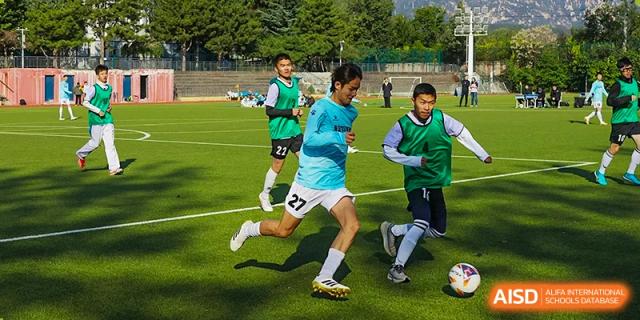 Two students in light blue and green jerseys compete for the soccer/football during a match or drill on the school field at Beijing Bayi School International Department.