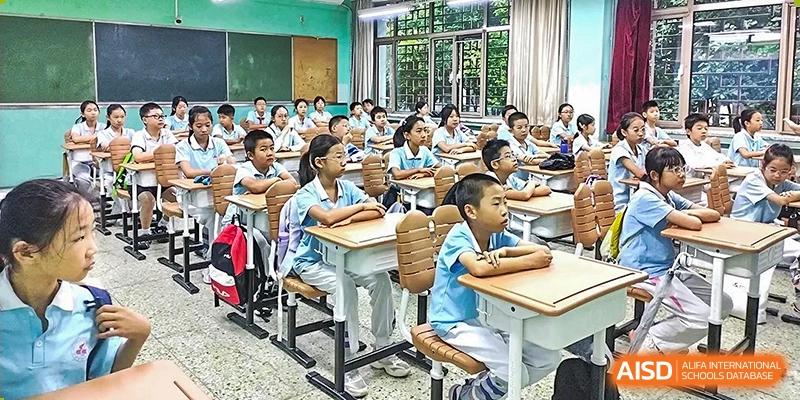 A busy classroom scene with multiple young students in uniforms seated at desks, listening or working, inside the Beijing Bayi School International Department.