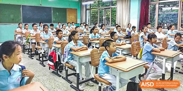 A busy classroom scene with multiple young students in uniforms seated at desks, listening or working, inside the Beijing Bayi School International Department.