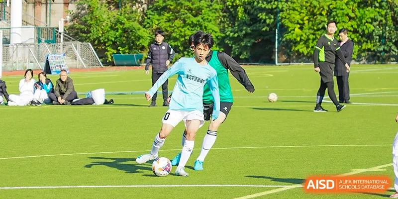 A student from the Beijing Bayi School International Department practices soccer/football on a green field, dribbling the ball past a teammate during a training session.