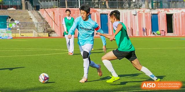 Intense action shot of two students, one in a light blue jersey and one in a green jersey, battling for possession of the soccer/football on the pitch at Beijing Bayi School International Department.